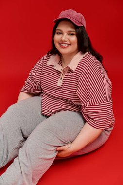 Güzel, iri yarı bir kadın, parlak kırmızı bir arka planda çizgili bir elbiseyle rahatlar..