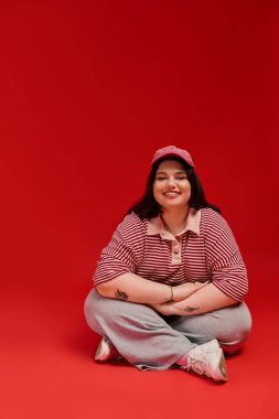 Büyük beden bir model, neşeli çizgili bir elbiseyle canlı kırmızı bir arka plana gülümsüyor..