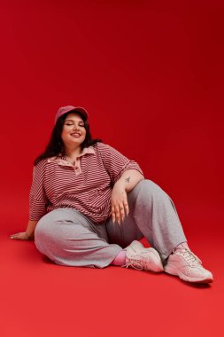 Parlak kırmızı arka plan, modaya uygun kıyafetini gösteren ve gülümseyen iri yarı bir kadın için zemin hazırlıyor..