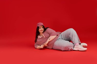 Ayrıca cüsseli bir kadın, koyu kırmızı bir zemine karşı kendine güvenerek poz veriyor..