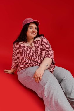 Modaya uygun ve cüsseli bir kadın kendine güvenen kırmızı bir yüzey üzerinde poz veriyor, eşsiz tarzını sergiliyor..