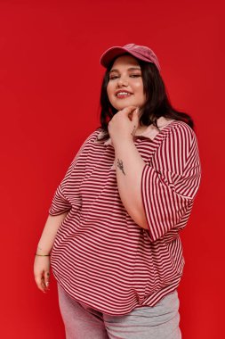 Güzel, iri yarı bir kadın, parlak kırmızı bir duvara şakacı bir şekilde poz veriyor. Moda kıyafetlerini sergiliyor..