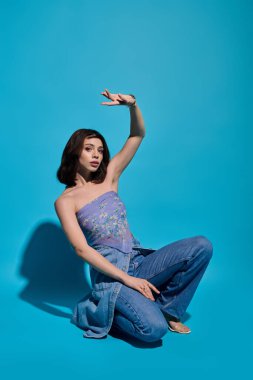 Güzel, genç bir kadın zarif bir şekilde diz çöker, çarpıcı bir ortamda modaya uygun kıyafetlerini sergiler..