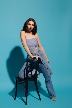 Çekici genç kadın kendine güvenen bir şekilde bir sandalyede poz veriyor. Benzersiz moda anlayışını ve güzelliğini vurguluyor..