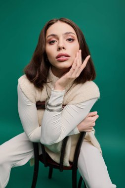 Şık genç bir kadın zarif bir şekilde bir sandalyede poz veriyor, yeşil bir zemine karşı kıyafetini sergiliyor..