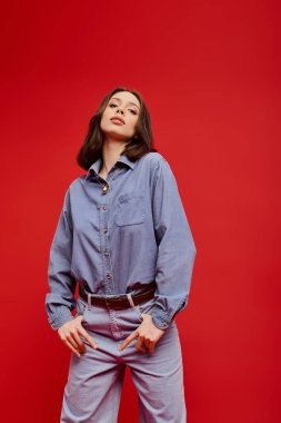 Genç bir kadın, koyu kırmızı arka planda şık bir kotla güven içinde poz veriyor..