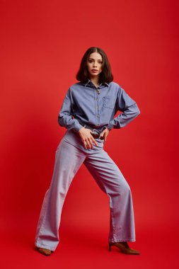 Şık bir kadın koyu kırmızı bir zemine karşı kot pantolonla rahatça poz veriyor..