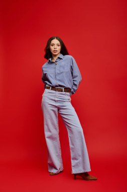 Genç bir kadın şık bir kot giysisi sergiliyor, koyu kırmızı bir arka plana karşı, zarafet saçıyor..