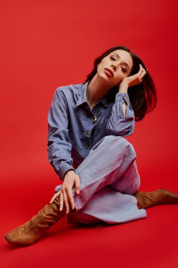 Şık bir genç kadın, parlak kırmızı bir arka planda özgüven ve moda yayarak düşünceli bir şekilde oturur..