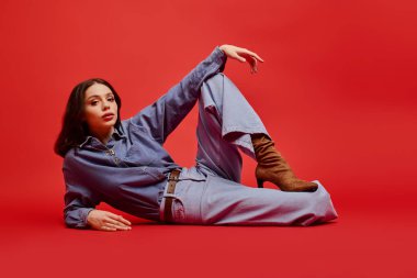 Genç bir kadın şık elbisesini ve botlarını sergileyerek kırmızı bir zeminde zarif bir poz veriyor..