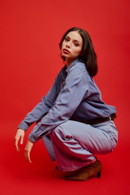 Modaya uygun genç bir kadın şık bir şekilde kot giyip, kırmızı bir arka plana yaslanıyor..