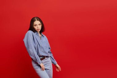 Şık kot elbiseli genç bir kadın zarif bir şekilde kırmızı bir zemine karşı duruyor..