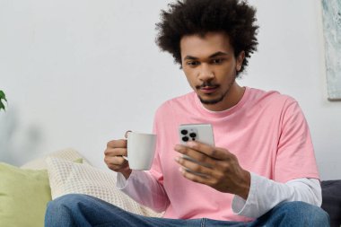 Pembe gömlekli şık bir genç adam kanepede rahatlar, çayını yudumlar ve telefonunu kaydırır..