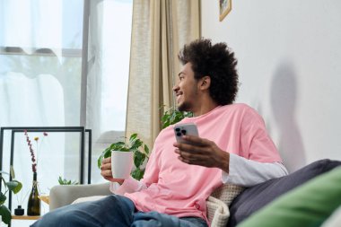 Güneşli bir yaşam alanında, mutlu genç bir adam bir fincan ve akıllı telefonla rahatlar..