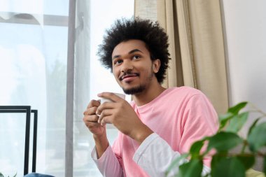 Gündelik giyinmiş genç bir adam pencerenin kenarında oturur, kahvenin tadını çıkarır ve yansıtır..