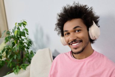 Kıvırcık saçlı gülümseyen genç adam bitkilerle dolu rahat bir odada dinlenirken kulaklık takıyor..
