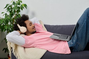 Yakışıklı genç bir adam, dizüstü bilgisayarını kullanırken kulaklık takarak kanepede uzanıyor..