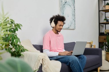 Genç adam kulaklık takıyor ve rahat bir koltukta otururken dizüstü bilgisayarına odaklanıyor..