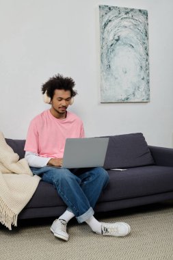 Gündelik giysiler içindeki yakışıklı genç adam rahat bir şekilde kanepede oturur ve dizüstü bilgisayarına odaklanır..