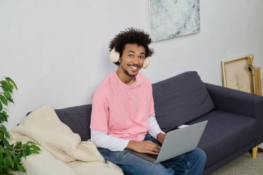Pembe tişörtlü neşeli genç bir adam laptopunu kullanır, sıcak ve davetkar bir oturma odasıyla çevrilidir..