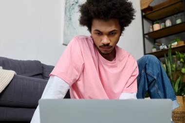 Yakışıklı genç adam yerde oturuyor, dizüstü bilgisayarına odaklanmış, bitkiler ve modern dekorlarla çevrili..