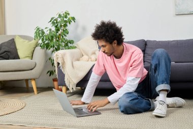 Odaklanmış genç bir adam halıya oturmuş, parlak, davetkar bir oturma odasında dizüstü bilgisayarla çalışıyor..