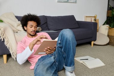 Genç bir adam rahat bir şekilde yerde oturur, kitap okumaya dalmış, rahat bir ortamda..