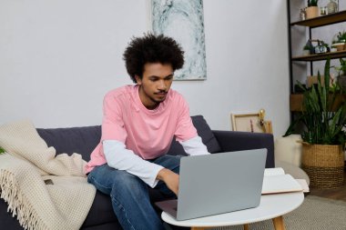 Genç adam rahat bir şekilde kanepede oturuyor, şık ve modern bir odada laptopuyla çalışıyor..