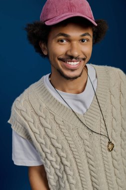 Çekici bir gülümsemesi olan genç bir adam, rahat örülmüş bir yelek ve şapka giyerken kendine güveniyor..