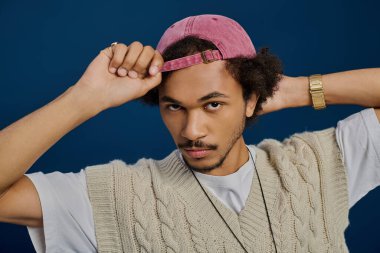 Kıvırcık saçlı, kendine güvenen genç adam şapkasını düzeltiyor, modaya uygun bir görünüm sergiliyor ve çarpıcı bir bakış sergiliyor..