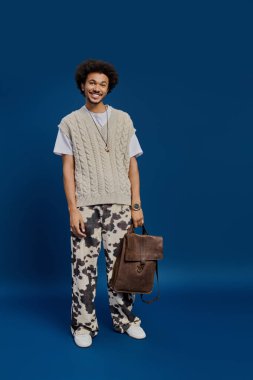 Kahverengi çantalı ve şık görünümlü genç bir adam mavi bir arka plana sıcak bir şekilde gülümsüyor..