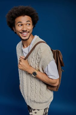 Kıvırcık saçlı yakışıklı genç adam neşeyle kahverengi bir sırt çantası taşıyor ve rahat bir kazak giyiyor.