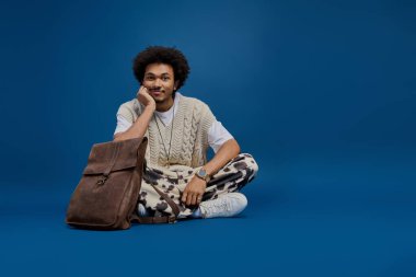 Çekici bir gülümsemesi olan genç bir adam parlak mavi bir zemine karşı bacak bacak üstüne atmış oturuyor..