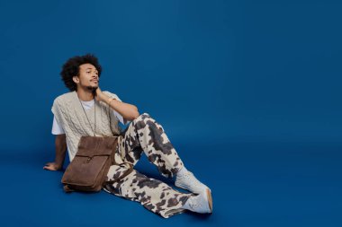 Genç bir adam yerde rahatça oturuyor, eşsiz tarzını ve rahat cazibesini sergiliyor..