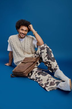 Genç bir adam mavi bir arkaplanda oturmuş neşeli bir gülümsemeyle canlı kıyafetini sergiliyor..