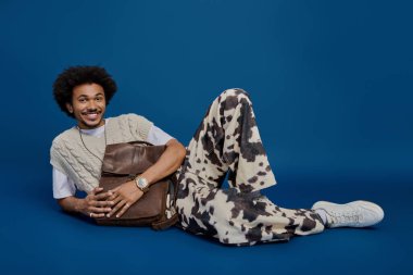 Genç bir adam parlak mavi bir arka planda rahat bir şekilde uzanır, şık kıyafetini ve çantasını sergiler..