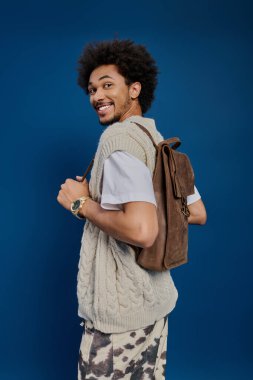 Parlak giyimli genç adam sıcak bir gülümsemeyle kendine güveni yayıyor, şık sırt çantasını gösteriyor..