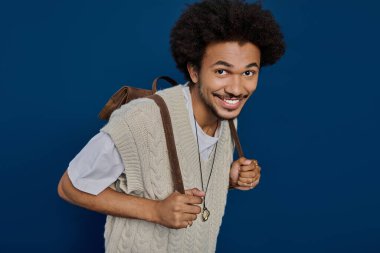 Örgü örmüş genç adam, parlak bir ortamda sırt çantasını tutarken neşeli bir şekilde ışıldıyor..