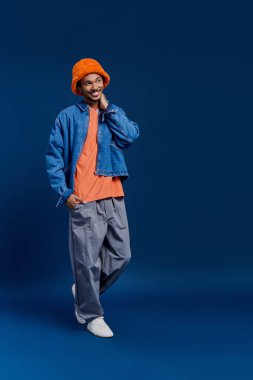 Yakışıklı genç adam kot ceket giyiyor, turuncu kova şapka giyiyor, rahat pantolon giyiyor, kendinden çok emin..