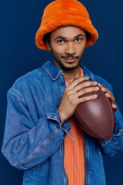 Turuncu şapkalı yakışıklı genç adam elinde futbol topu tutuyor, neşeli bir şekilde gülümsüyor..
