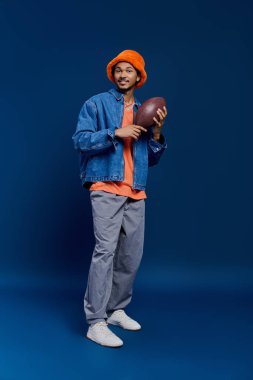 Yakışıklı genç bir adam futbol topu tutuyor, canlı renklerde modern bir kıyafet giyiyor, yaratıcı bir şekilde poz veriyor..