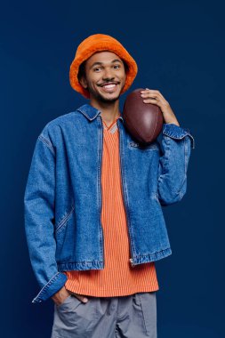 Genç ve yakışıklı bir adam gururla duruyor, canlı renkler giymiş, elinde bir futbol topu tutuyor..