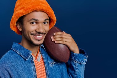 Turuncu şapkalı neşeli bir genç adam elinde bir futbol topuyla büyüleyici bir poz veriyor..