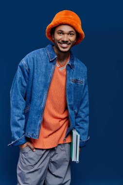 Kot ceket giyen ve turuncu şapkalı genç adam neşeli poz veriyor, kitapları sıradan bir şekilde tutuyor..