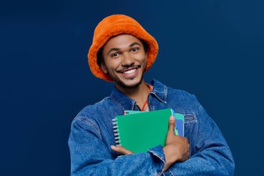 Neşeli bir gülüşü olan yakışıklı genç adam elinde renkli defterler, kendinden emin duruyor..