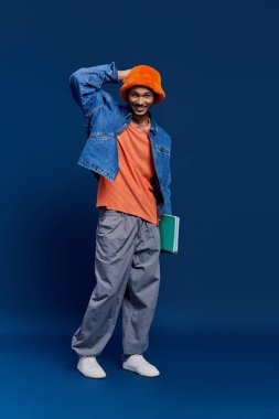 Turuncu şapkalı ve kot ceketli yakışıklı genç adam elinde yeşil bir kitapla rahatça poz veriyor..