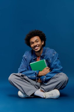Genç, şık, kıvırcık saçlı bir adam elinde yeşil bir defter, mavi bir arka planda gülümserken..