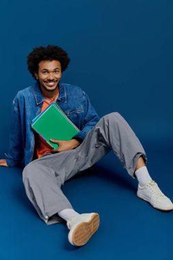 Genç bir adam yerde oturuyor, şık giysiler içinde, canlı bir not defteriyle gülümsüyor..