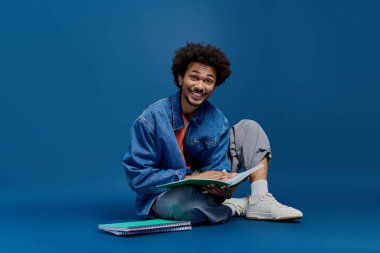 Gülümseyen genç adam yerde oturuyor, renkli defterlerle çevrili, çalışmayla meşgul..