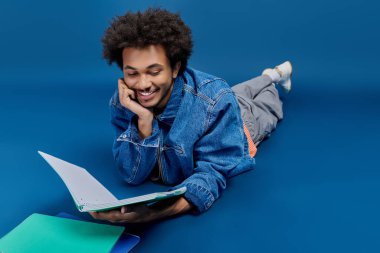 Genç, yakışıklı bir adam yerde yatıyor, renkli dosyalarla çevrili bir kitapla mutlu bir şekilde..